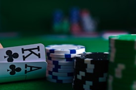 A casino scene featuring two playing cards, the ace and king of clubs, partially concealed by a player's hand. Nearby, there are stacks of multicolored poker chips on a green felt table, with blurred colorful background elements suggesting other chip stacks or cards.