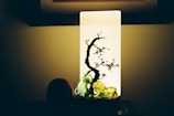 A decorative light featuring a rectangular panel with a silhouette of a tree and branches with small flowers. Below the light panel, there are colorful figurines, including a green caterpillar and other shapes. The background is dimly lit, enhancing the contrast between the illuminated panel and its surroundings.