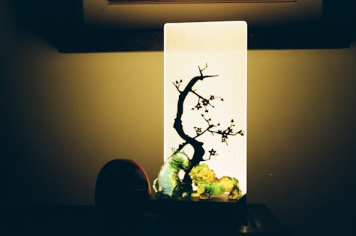 A decorative light featuring a rectangular panel with a silhouette of a tree and branches with small flowers. Below the light panel, there are colorful figurines, including a green caterpillar and other shapes. The background is dimly lit, enhancing the contrast between the illuminated panel and its surroundings.