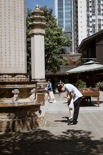 A man dressed in a light blue shirt and black pants bending down to assist a child in white, set in an outdoor courtyard area. The background features a stone pillar engraved with gold Chinese characters, additional architectural elements, some trees, and a high-rise building. Two other individuals are seen walking and standing in the background.