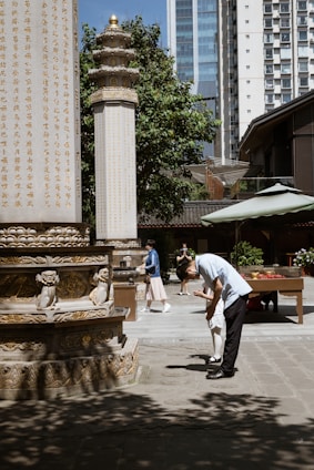 A man dressed in a light blue shirt and black pants bending down to assist a child in white, set in an outdoor courtyard area. The background features a stone pillar engraved with gold Chinese characters, additional architectural elements, some trees, and a high-rise building. Two other individuals are seen walking and standing in the background.
