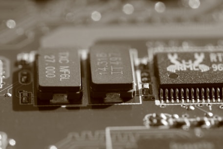 A close-up view of an electronic circuit board, featuring several visible electronic components such as integrated circuits and capacitors. The image has a sepia tone, giving it a vintage appearance, and the components are detailed with numbers and letters etched on their surfaces.