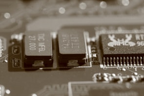 A close-up view of an electronic circuit board, featuring several visible electronic components such as integrated circuits and capacitors. The image has a sepia tone, giving it a vintage appearance, and the components are detailed with numbers and letters etched on their surfaces.