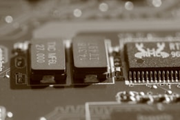 A close-up view of an electronic circuit board, featuring several visible electronic components such as integrated circuits and capacitors. The image has a sepia tone, giving it a vintage appearance, and the components are detailed with numbers and letters etched on their surfaces.