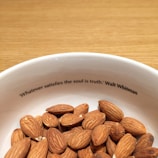 A white bowl filled with almonds sits on a wooden surface. Inside the rim of the bowl is a printed quote: 'Whatever satisfies the soul is truth.' by Walt Whitman.