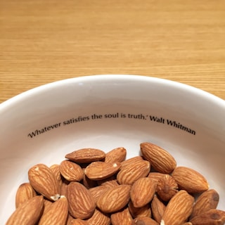 A white bowl filled with almonds sits on a wooden surface. Inside the rim of the bowl is a printed quote: 'Whatever satisfies the soul is truth.' by Walt Whitman.