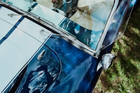 A close-up view of a vintage car, showcasing its glossy blue and white exterior. The reflection of tree branches and the sky can be seen on the car's surface. The photograph includes details like a side mirror, windshield wipers, and part of the interior with cream-colored upholstery.