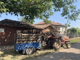 A rural scene featuring a red tractor attached to a blue trailer loaded with crates. The setting includes a rustic house with a tiled roof and a stack of wooden logs. Tall trees create a shaded area, and the sky is clear, suggesting a bright day.