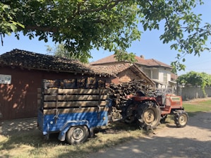 A rural scene featuring a red tractor attached to a blue trailer loaded with crates. The setting includes a rustic house with a tiled roof and a stack of wooden logs. Tall trees create a shaded area, and the sky is clear, suggesting a bright day.