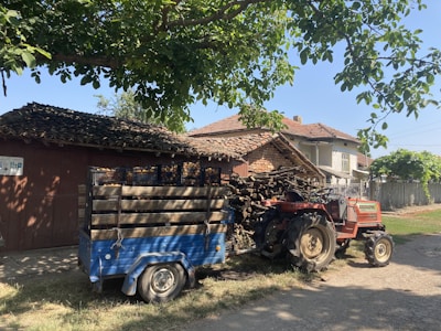 A rural scene featuring a red tractor attached to a blue trailer loaded with crates. The setting includes a rustic house with a tiled roof and a stack of wooden logs. Tall trees create a shaded area, and the sky is clear, suggesting a bright day.