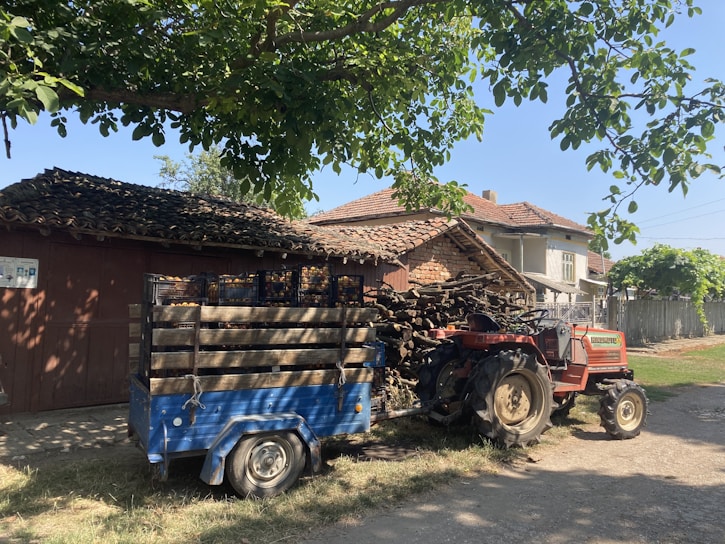A rural scene featuring a red tractor attached to a blue trailer loaded with crates. The setting includes a rustic house with a tiled roof and a stack of wooden logs. Tall trees create a shaded area, and the sky is clear, suggesting a bright day.