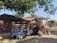 A rural scene featuring a red tractor attached to a blue trailer loaded with crates. The setting includes a rustic house with a tiled roof and a stack of wooden logs. Tall trees create a shaded area, and the sky is clear, suggesting a bright day.