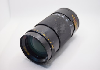 A black camera lens with various measurement markings in yellow and white, featuring a textured grip for manual adjustment. The lens is positioned at an angle on a light surface.