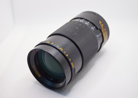 A black camera lens with various measurement markings in yellow and white, featuring a textured grip for manual adjustment. The lens is positioned at an angle on a light surface.