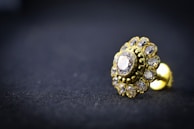 A close-up of a gold-toned earring with a central diamond surrounded by smaller gemstones, resting on a dark textured surface. The intricate design is highlighted by the shiny metal and transparent stones, creating a luxurious and elegant appearance.
