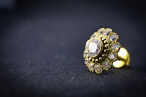 A close-up of a gold-toned earring with a central diamond surrounded by smaller gemstones, resting on a dark textured surface. The intricate design is highlighted by the shiny metal and transparent stones, creating a luxurious and elegant appearance.