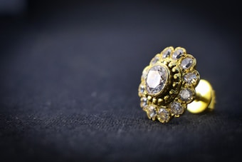 A close-up of a gold-toned earring with a central diamond surrounded by smaller gemstones, resting on a dark textured surface. The intricate design is highlighted by the shiny metal and transparent stones, creating a luxurious and elegant appearance.