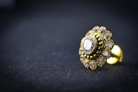 A close-up of a gold-toned earring with a central diamond surrounded by smaller gemstones, resting on a dark textured surface. The intricate design is highlighted by the shiny metal and transparent stones, creating a luxurious and elegant appearance.