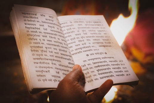 A person's hand holding an open book written in an ancient script, possibly Sanskrit. The background shows a warm, glowing fire, suggesting a ritualistic or spiritual setting.