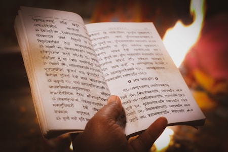 A person's hand holding an open book written in an ancient script, possibly Sanskrit. The background shows a warm, glowing fire, suggesting a ritualistic or spiritual setting.
