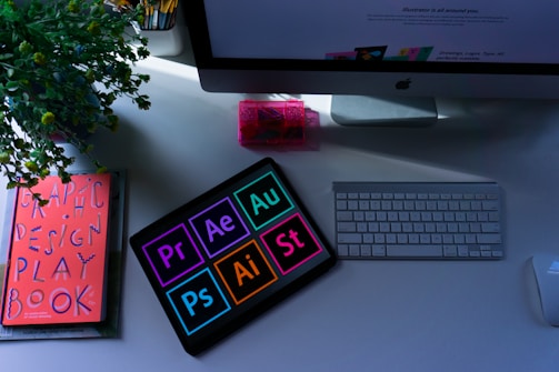 A workspace featuring a keyboard, mouse, monitor, and a tablet displaying icons for design software such as Photoshop, Premiere, After Effects, Illustrator, and Audition. A book titled 'Graphic Design Play Book' is positioned next to a plant, and a pink transparent container is near the monitor.