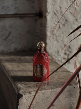 A red bottle with a gold cap stands on a rustic stone surface, with intricate detailing and a label featuring ornate designs in the center. The scene is partially shadowed, suggesting sunlight filtering through nearby foliage, which is partially visible on the right.