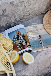 An open fashion magazine lies on a textured stone surface, displaying images of a model in outdoor settings. Beside the magazine, a clear glass of water rests next to a straw sun hat. A reusable net bag containing lemons and two lemon halves are also visible.
