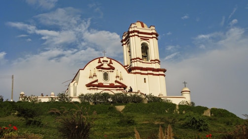 A white church with red trim sits atop a grassy hill under a blue sky with scattered clouds. The building features a tall bell tower and several architectural details like arches and a cross. Vegetation surrounds the church, and a few people are seen near the entrance.