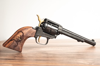 A detailed image of a revolver with a long barrel resting on a wooden surface. The handle is made of polished wood featuring an engraved scorpion design. The revolver has metallic components with a combination of black and gold accents.