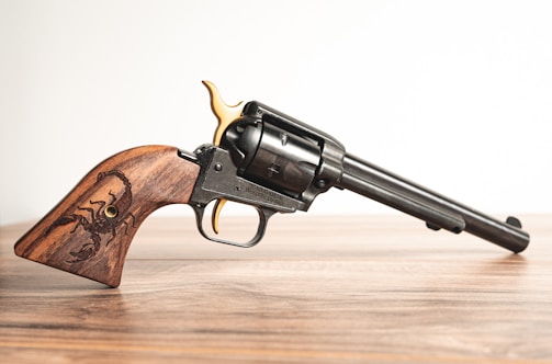 A detailed image of a revolver with a long barrel resting on a wooden surface. The handle is made of polished wood featuring an engraved scorpion design. The revolver has metallic components with a combination of black and gold accents.