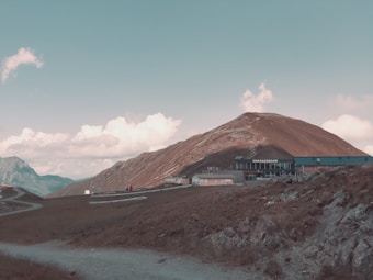 A mountainous landscape with a clear blue sky and scattered clouds. A brown hill rises prominently in the center with a building labeled 'SMARAGDBAHN' situated at the base. The foreground includes a winding gravel path and rocky terrain, with distant mountain peaks visible on the left.