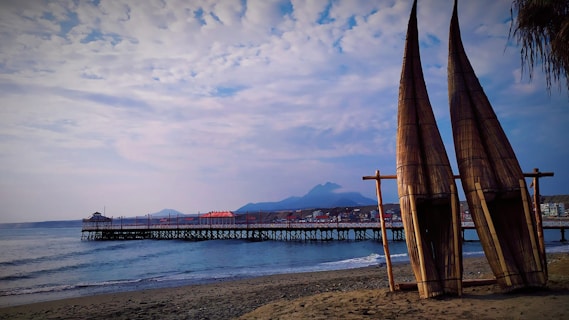 Two traditional reed boats are resting on a sandy beach with a calm ocean stretching out to a wooden pier. In the background, mountains rise against a partly cloudy sky, while small buildings line the shore.