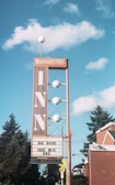 A vintage-style inn sign stands against a blue sky with fluffy clouds. The sign is tall with a vertical 'INN' text and has circular lights along its structure. Below, a marquee advertises room rates, free WiFi, and HBO, with a 'No Vacancy' notice. Surrounding the sign are evergreen trees and a red brick building with green roof accents.