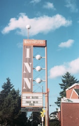 A vintage-style inn sign stands against a blue sky with fluffy clouds. The sign is tall with a vertical 'INN' text and has circular lights along its structure. Below, a marquee advertises room rates, free WiFi, and HBO, with a 'No Vacancy' notice. Surrounding the sign are evergreen trees and a red brick building with green roof accents.