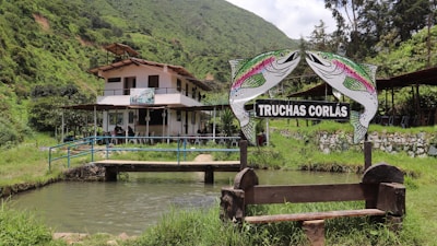 A building with a balcony and patio is nestled in a lush green mountainous area. In the foreground, there is a sign featuring two fish and the text 'Truchas Corlas' above a small pond. The surrounding area is filled with greenery and trees.