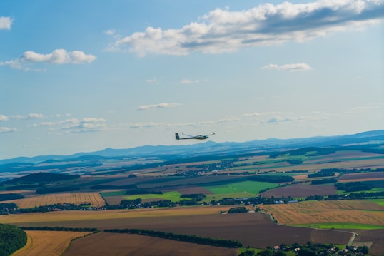 A glider is flying gracefully over an expansive rural landscape characterized by patchwork fields and gentle rolling hills. The horizon is marked by distant mountain ranges under a clear blue sky dotted with scattered clouds.
