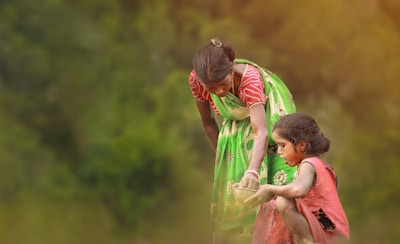 A woman in a colorful sari is crouching down and holding a bowl as a young girl, covered in dust or paint, balances nearby. They are outdoors with a blurred green background, which suggests a rural setting.