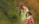 A woman in a colorful sari is crouching down and holding a bowl as a young girl, covered in dust or paint, balances nearby. They are outdoors with a blurred green background, which suggests a rural setting.