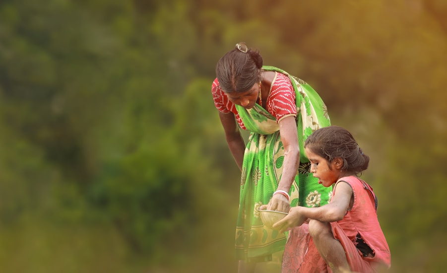 A woman in a colorful sari is crouching down and holding a bowl as a young girl, covered in dust or paint, balances nearby. They are outdoors with a blurred green background, which suggests a rural setting.