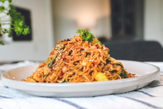 A plate of stir-fried noodles garnished with sesame seeds and fresh herbs, possibly parsley. The noodles appear to be mixed with vegetables and small pieces of meat, and the dish is presented in an appetizing manner.