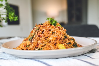 A plate of stir-fried noodles garnished with sesame seeds and fresh herbs, possibly parsley. The noodles appear to be mixed with vegetables and small pieces of meat, and the dish is presented in an appetizing manner.