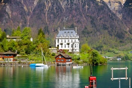 A grand, elegant mansion with a classic architectural style is surrounded by lush greenery and set against a backdrop of towering mountains. In the foreground, a serene lake reflects the buildings and the natural surroundings, with a couple of small boats docked by a wooden pier. A quaint wooden house is also visible near the shoreline, adding to the tranquil and picturesque setting.