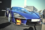 A sleek blue high-performance McLaren sports car is prominently displayed. The vehicle features sharp lines and aerodynamic contours, with visible carbon fiber elements and distinctive headlights. It is parked on a black surface, with other cars and people visible in the background.