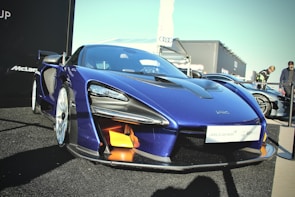 A sleek blue high-performance McLaren sports car is prominently displayed. The vehicle features sharp lines and aerodynamic contours, with visible carbon fiber elements and distinctive headlights. It is parked on a black surface, with other cars and people visible in the background.