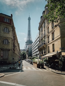 A street scene featuring the Eiffel Tower in the background, surrounded by classic Parisian architecture. The street is lined with apartment buildings and a few cafes with outdoor seating. Trees partially shade the sidewalk, and there are few people visible, creating a calm urban atmosphere.