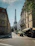 A street scene featuring the Eiffel Tower in the background, surrounded by classic Parisian architecture. The street is lined with apartment buildings and a few cafes with outdoor seating. Trees partially shade the sidewalk, and there are few people visible, creating a calm urban atmosphere.