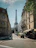 A street scene featuring the Eiffel Tower in the background, surrounded by classic Parisian architecture. The street is lined with apartment buildings and a few cafes with outdoor seating. Trees partially shade the sidewalk, and there are few people visible, creating a calm urban atmosphere.