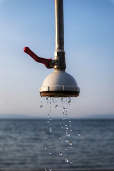A metal showerhead is mounted outdoors with water droplets flowing down against the backdrop of a calm sea and a clear sky. The shower pipe is metallic with a red handle attached.