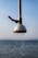 A metal showerhead is mounted outdoors with water droplets flowing down against the backdrop of a calm sea and a clear sky. The shower pipe is metallic with a red handle attached.