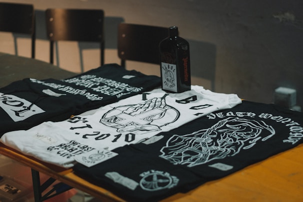 Three black and white graphic t-shirts are laid out on a wooden surface, featuring intricate designs and texts. A dark-colored square bottle, possibly an alcoholic beverage, is placed amongst the shirts. In the background, there are some unoccupied chairs with a dimly lit atmosphere surrounding the scene.